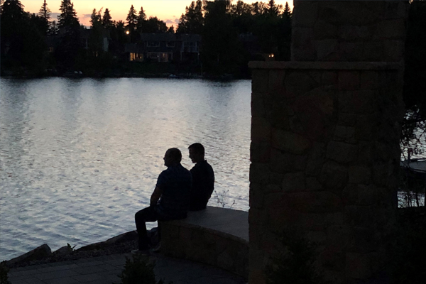 Two silhouetted figures sit on a stone bench by a calm lake at dusk, with soft colors reflecting on the water.