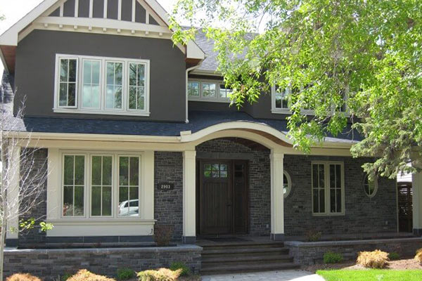 Front view of a modern, two-story farmhouse with a stone base, large windows, and a welcoming entrance surrounded by lush greenery.