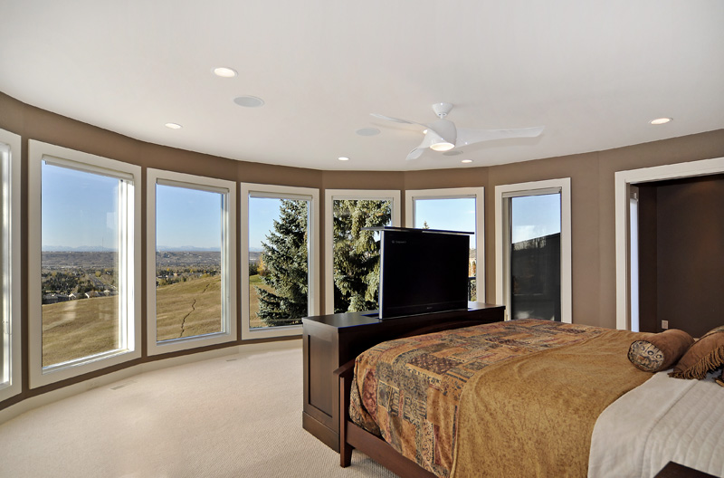Bright, modern bedroom with panoramic windows, a ceiling fan, and a flat-screen TV on a dark wood dresser, showcasing scenic views.