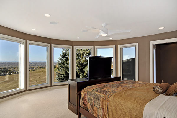 Bright, modern bedroom with panoramic windows, a ceiling fan, and a flat-screen TV on a dark wood dresser, showcasing scenic views.