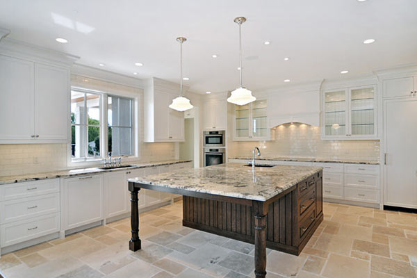 Bright, modern kitchen with white cabinetry, a large granite island, and pendant lighting, featuring natural stone flooring and appliances.