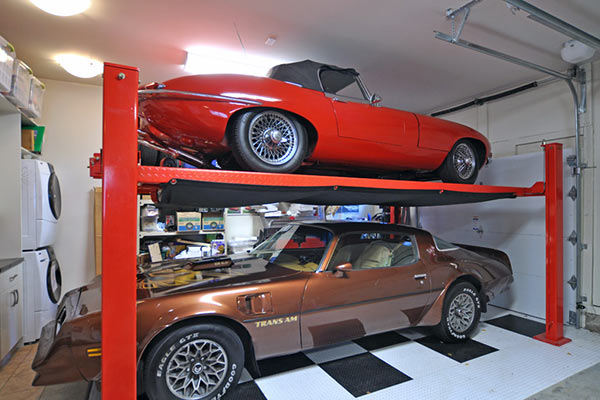 A red convertible sports car is elevated on a lift above a brown Trans Am in a tidy garage, surrounded by storage and laundry appliances.