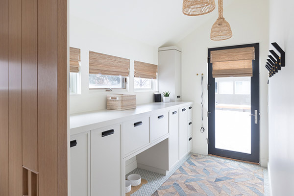 Bright, modern vestibule with white cabinetry, woven shades, and a black door, featuring a textured rug and organized pet bowls.