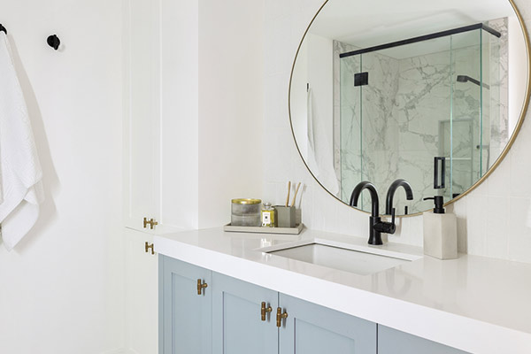 Modern bathroom featuring a round mirror, light blue cabinets, brass accents, and a sleek glass shower with marble walls.
