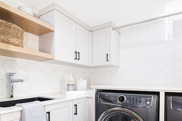Bright laundry room featuring white hexagon tile, sleek cabinetry, a modern washing machine, and natural wooden shelving. Inside custom home built by Cortina in Calgary, Alberta