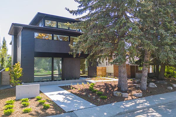 Contemporary custom Calgary home, surrounded by greenery, featuring large windows and a landscaped yard with trees and stone accents.