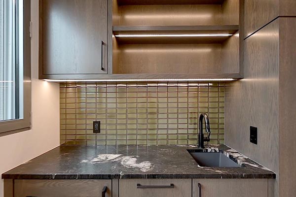 custom wet bar featuring a black marble countertop, decorative tile backsplash, sleek faucet, and wooden cabinets with LED lighting.