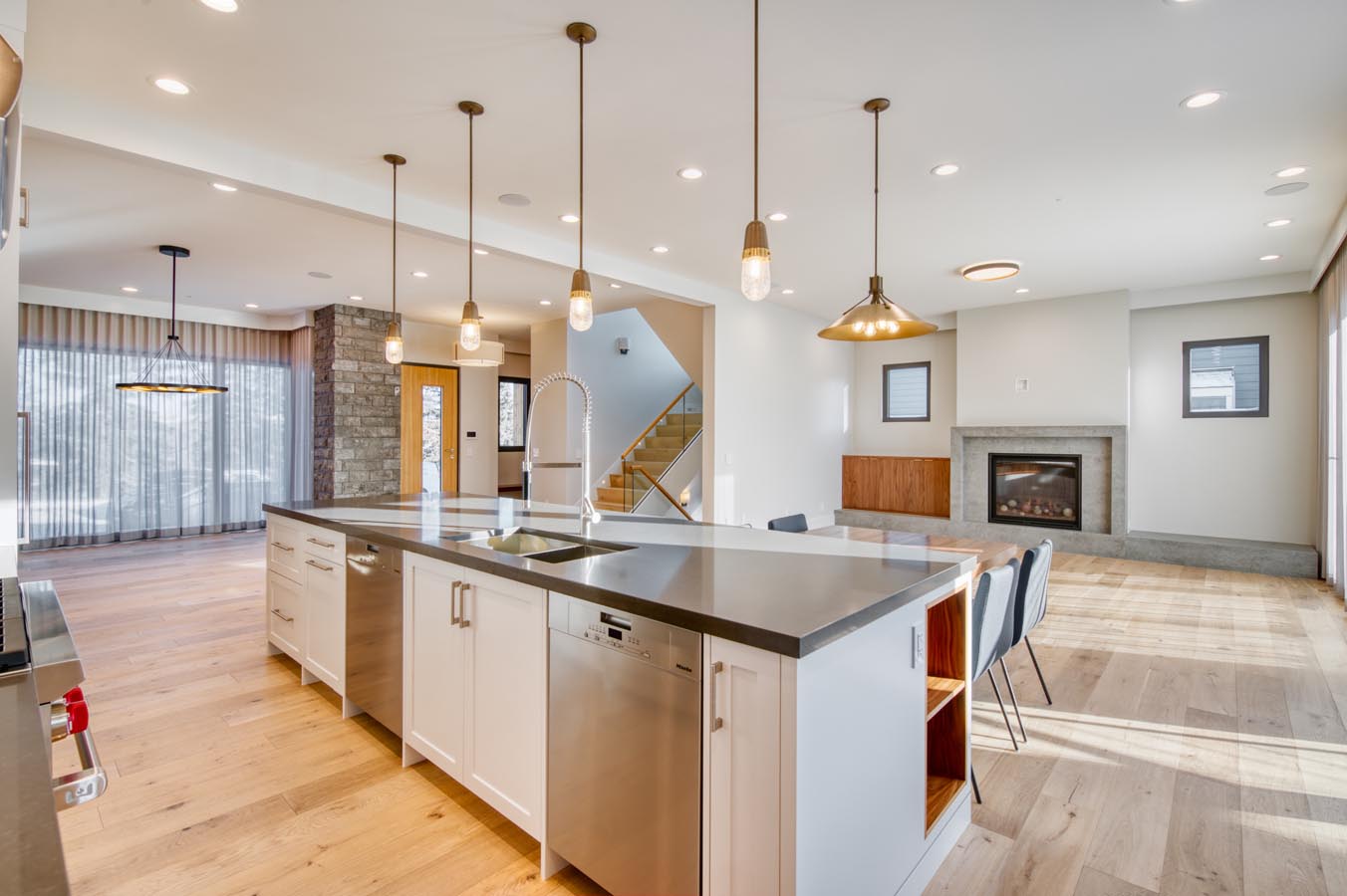 Modern kitchen with an island, elegant lighting, stainless appliances, and a cozy living area featuring a fireplace and wooden accents in custom home built by Cortina Crafted Homes